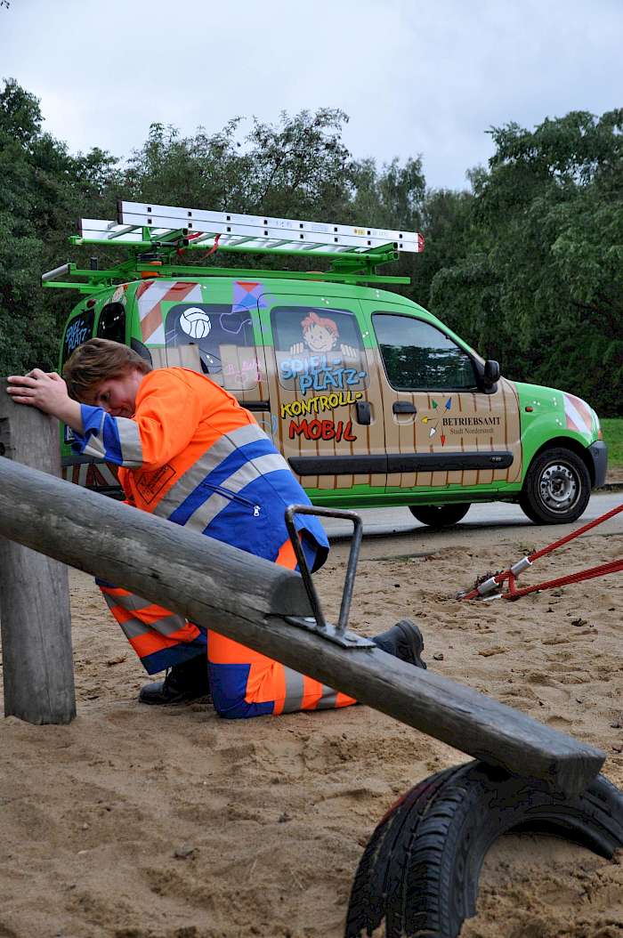 Spielplatzprüferin an Wippe. Im Hintergrund Spielplatzkrontrollfahrzeug